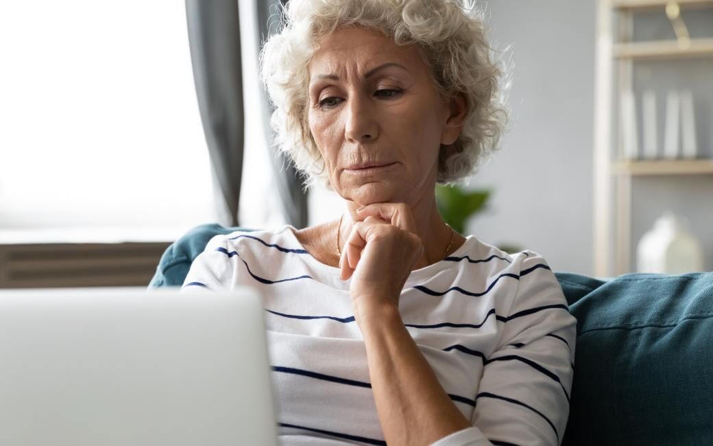 Een oudere vrouw zit nadenkend op de bank achter een laptop